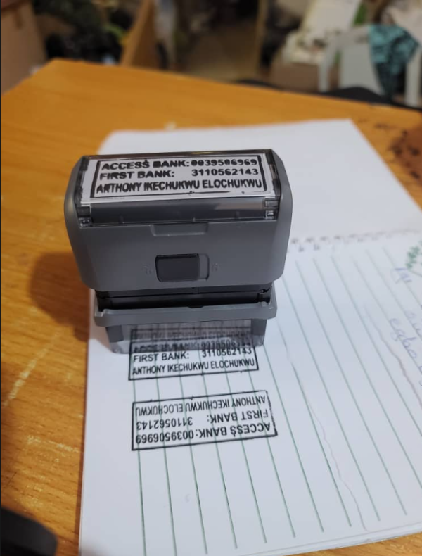 Self-inking rubber stamp showing Access Bank, First Bank, and account details, resting on stamped paper on a wooden desk
