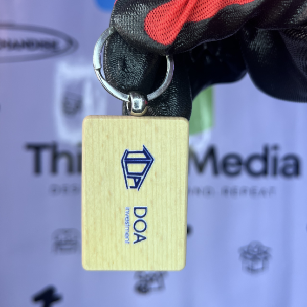 Wooden rectangular keychain with a blue diamond logo and "DOA" text, held against a Thimze Media branded backdrop